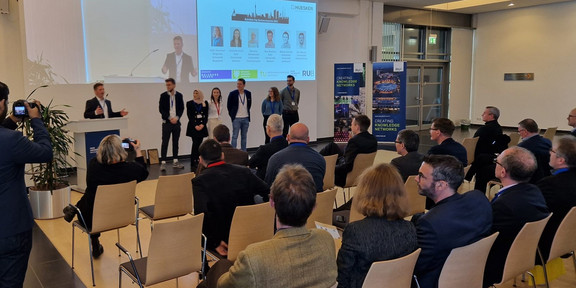 Auf dem Foto ist eine Konferenzsituation zu sehen. Mehrere Menschen befinden sich im vorderen Bereich, wo auch eine Beamerprojektion an der Wand zu sehen ist. Ein Redner stellt 5 Personen vor. Diesen werden von ca 20 sitzenden Personen zugehört.